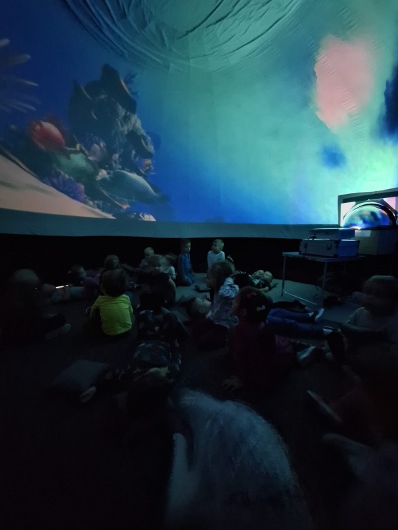 Dzieci siedzą lub leżą w mobilnym kinie sferycznym. W tle widać kolorowe ryby w oceanie.
