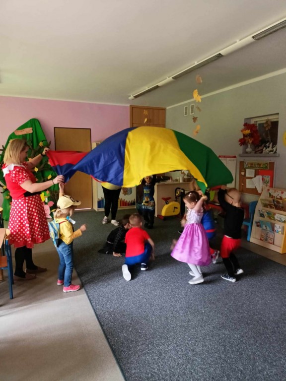 Dzieci wraz z nauczycielką stoją w kole i unoszą kolorową chustę do góry