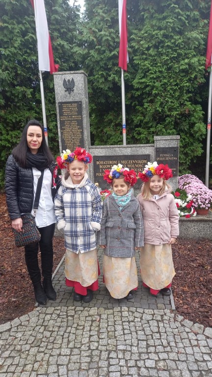 Dziewczynki z wiankami na głowie stoją przed pomnikiem z okazji Swieta Niepodleglosci razem ze swoja wychowawczynia