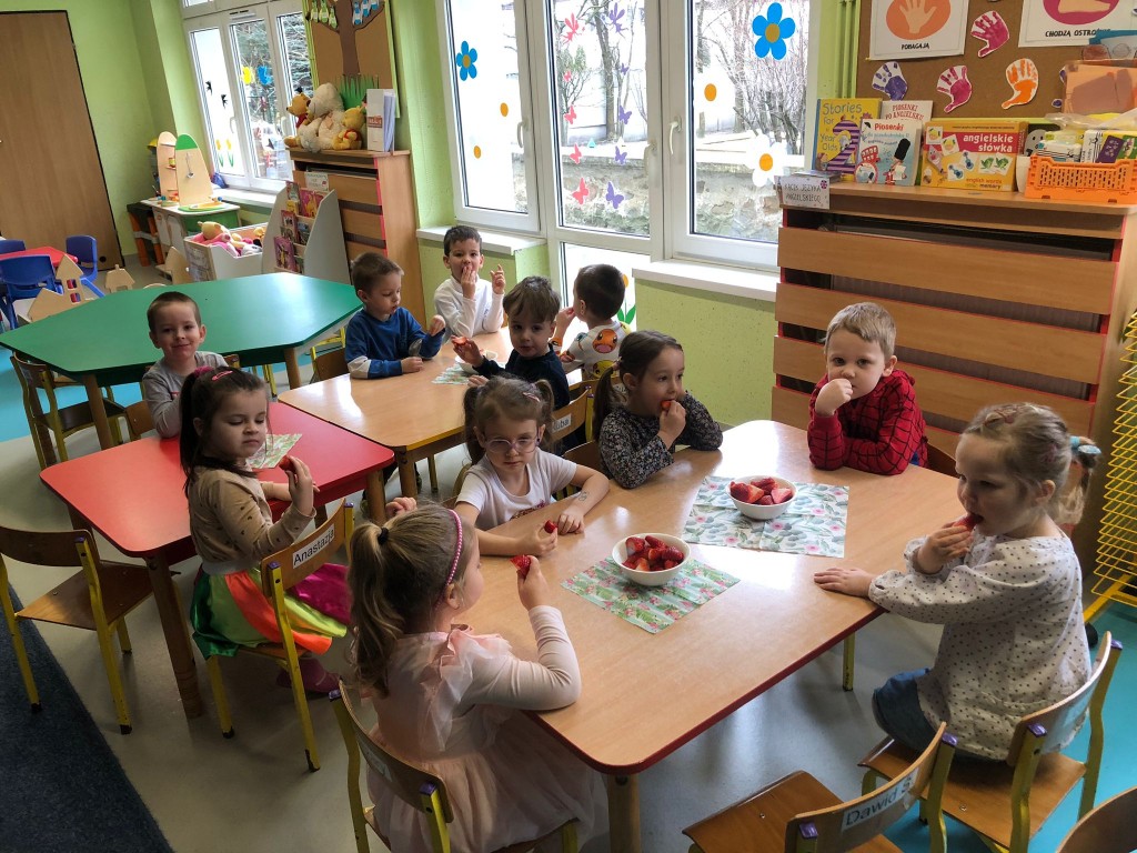 Dzieci siedzą przy stolikach i częstują się truskawkami