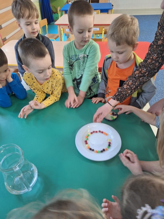 Nauczycielka pokazuje dzieciom jak zrobić tęczę z kolorowych drażetek