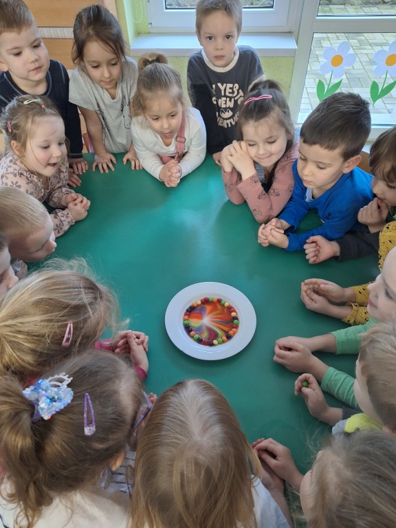 Dzieci stoją wokół stołu i obserwują doświadczenie z tęczą na talerzu