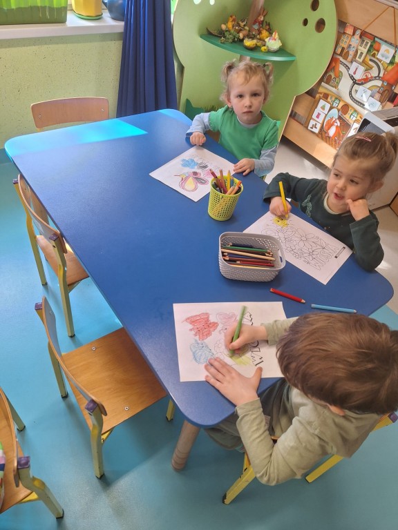 Dzieci siedzą przy stolikach i kolorują wiosenne obrazki