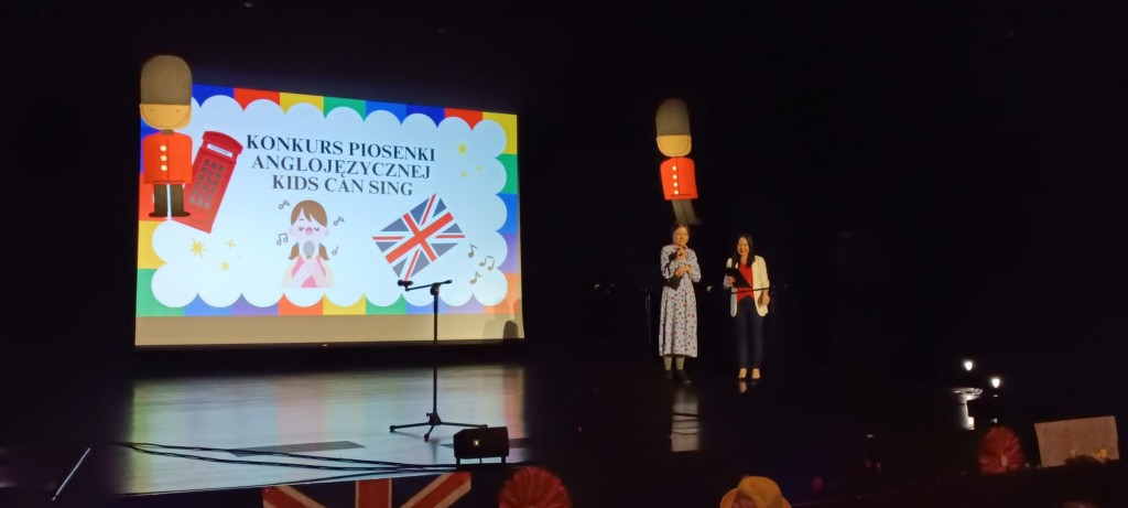dwie nauczycielki stoja na scenie i witaja dzieci na konkursie piosenki angielskiej