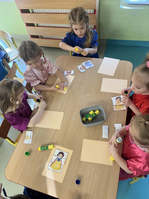 dziewczynki przy stoliku układają trzyelementowe puzzle z księżniczkami i naklejają na kartkę